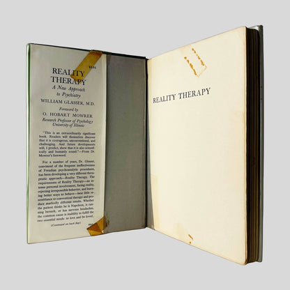 Inside front cover view of Reality Therapy by William Glasser, M.D. centered on a neutral light gray background. Dust jacket is reinforced to stay on the book with tape that has aged. 