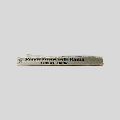 Close-up spine view of Rendezvous With Rama by Arthur C. Clarke centered on a neutral light gray background. Spine shows Chipping and creasing. Pages lay flat.