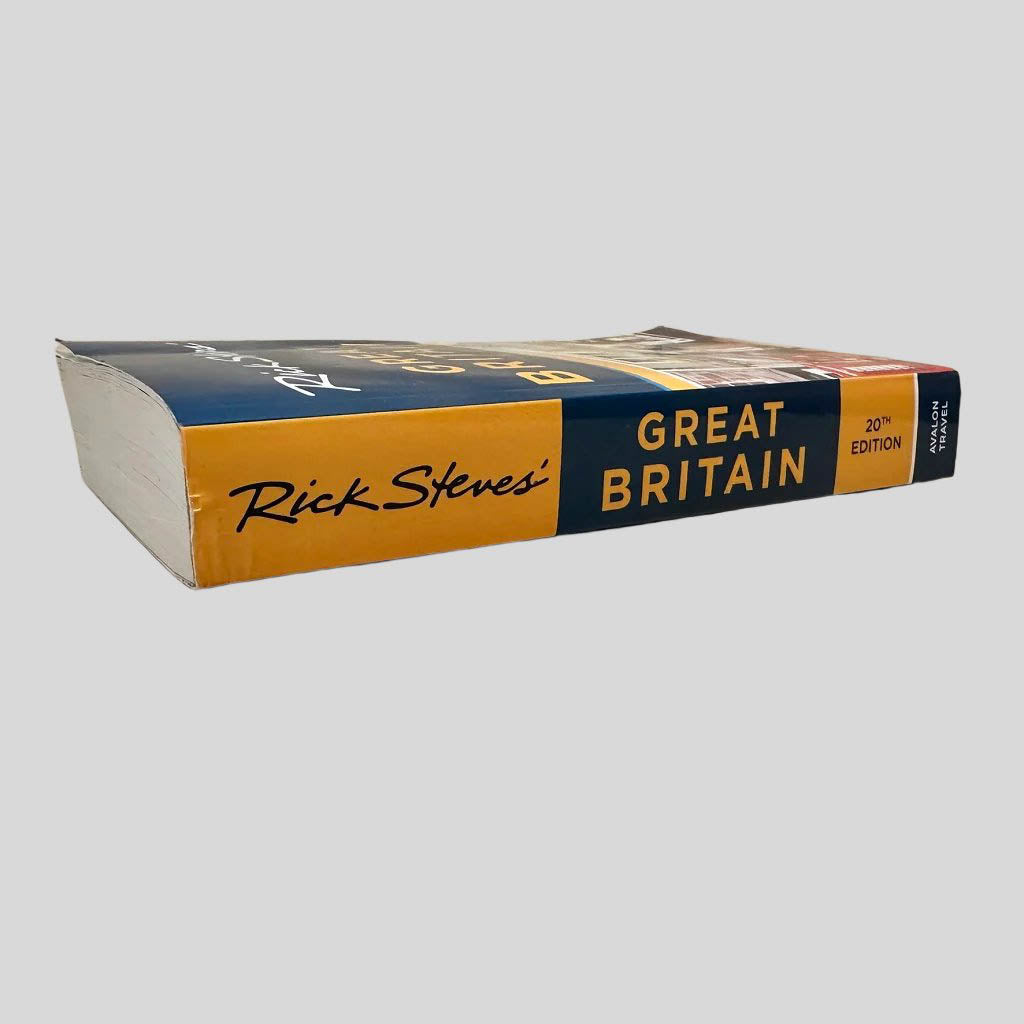 Close-up spine view of Rick Steves Great Britain 20th Edition centered on a neutral light gray background. Spine is square and shows creasing only at the top of the spine as a result of bumping. Pages lay flat.