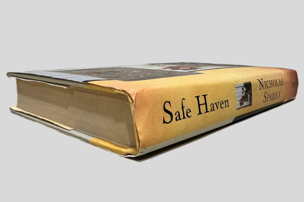 Close-up spine view of Safe Haven by Nicholas Sparks centered on a neutral light gray background. Dust jacket is present. Creasing at the top of the dust jacket. Pages lay flat.