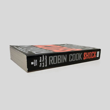 Close-up spine view of Shock by Robin Cook centered on a neutral light gray background. Spine is square and shows minimal creasing.