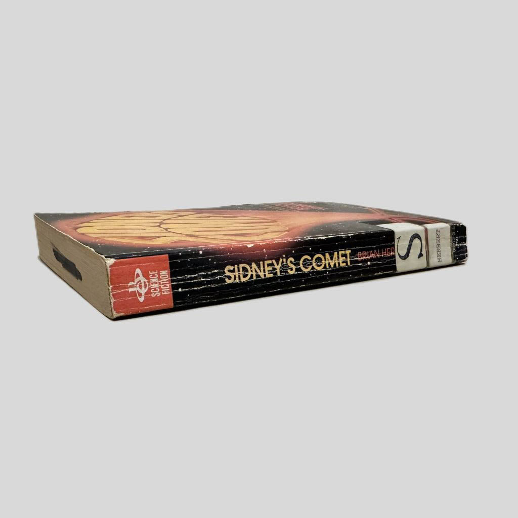 Close-up spine view of Sidney's Comet by Brian Herbert centered on a neutral light gray background. Spine shows creasing and chipping on the ends from wear. Library catalog stickers are taped at the bottom of the spine. Pages lay flat.