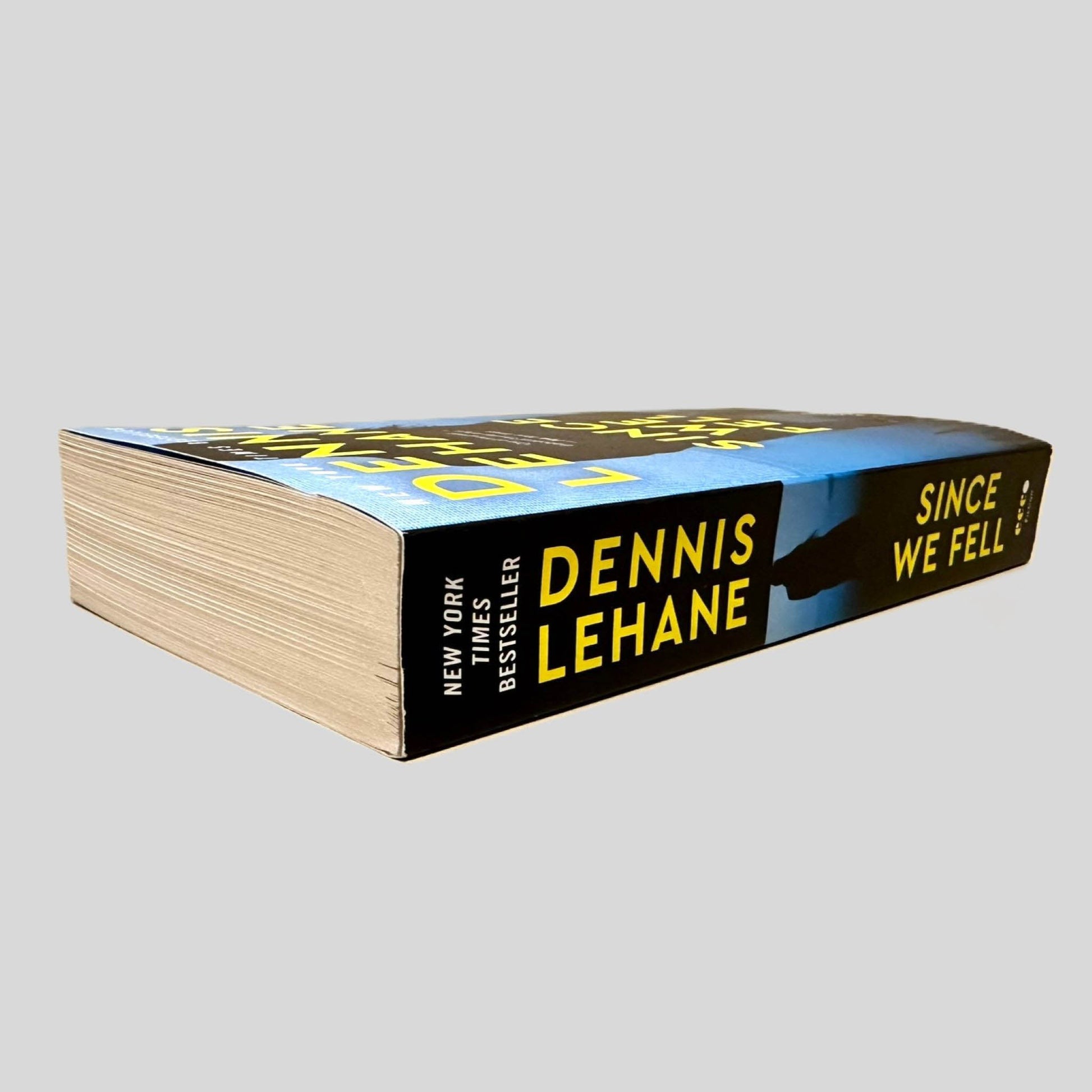 Close-up spine view of Since We Fell by Dennis Lehane centered on a neutral light gray background. Spine is square, shows no creasing. Pages lay flat.