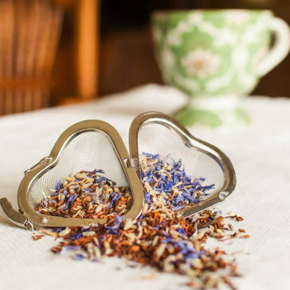 An open heart-shaped stainless steel mesh tea infuser containing loose-leaf herbal tea with blue cornflower petals, shown with a blurred green patterned teacup in the background.