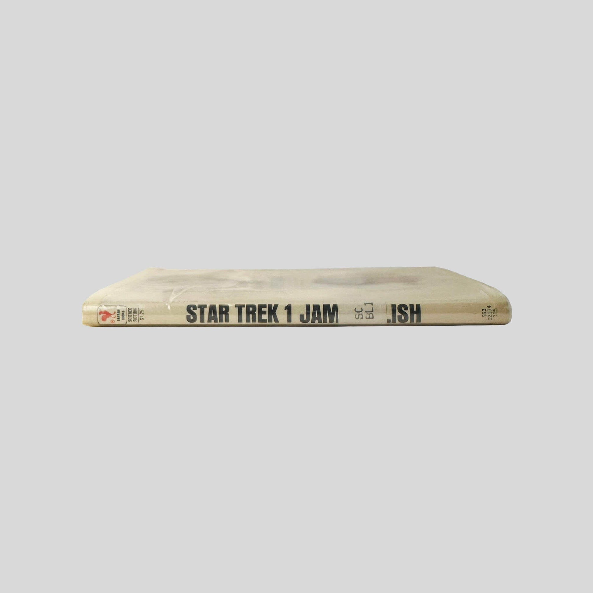 Close-up spine view of Star Trek 1 adapted by James Blish centered on a neutral light gray background. Spine shows creasing to the mylar cover and library shelving sticker is attached at the lower end of the spine. Pages lay flat.