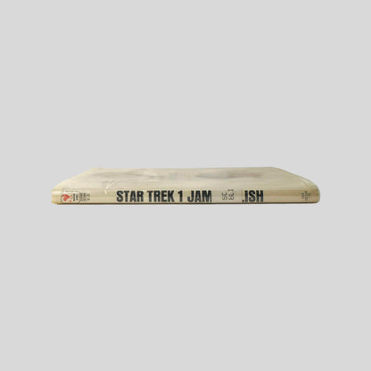 Close-up spine view of Star Trek 1 adapted by James Blish centered on a neutral light gray background. Spine shows creasing to the mylar cover and library shelving sticker is attached at the lower end of the spine. Pages lay flat.