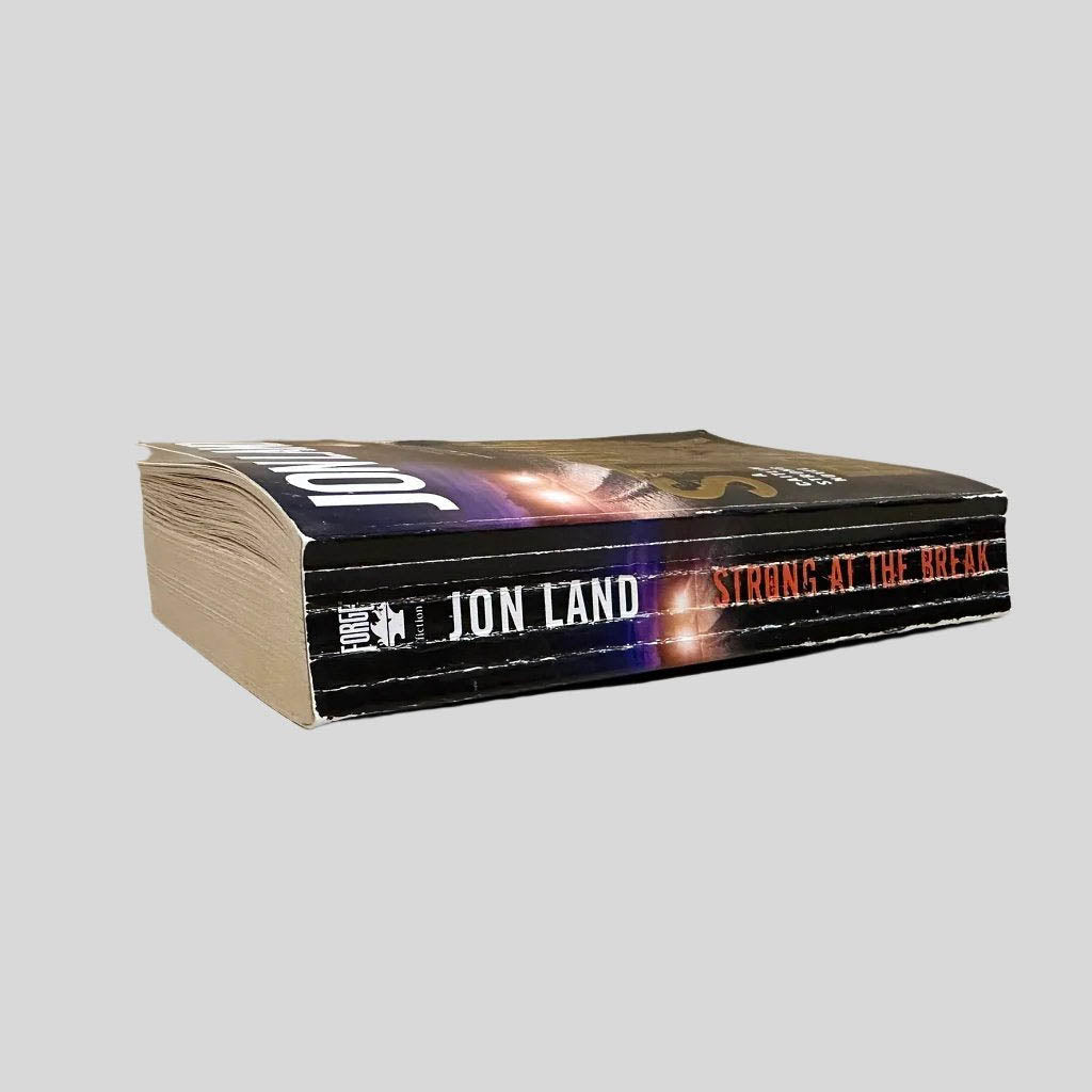 Close-up spine view of Strong at the Break by Jon Land centered on a neutral light gray background. Spine shows creasing. Top page edges show water damage, causing a slight wave for the top third of the book's pages. Text is in tact.