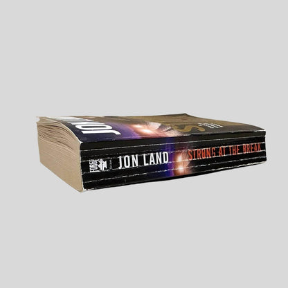 Close-up spine view of Strong at the Break by Jon Land centered on a neutral light gray background. Spine shows creasing. Top page edges show water damage, causing a slight wave for the top third of the book's pages. Text is in tact.