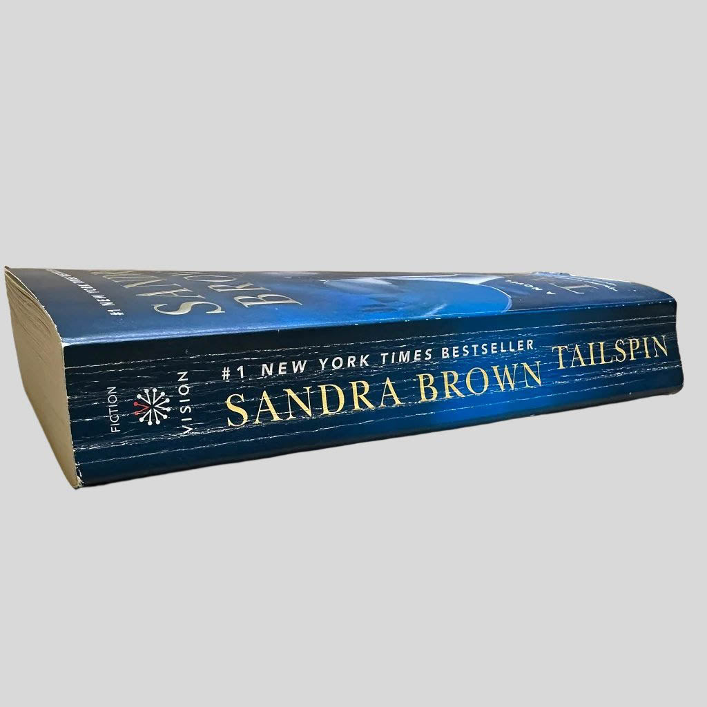Close-up spine view of Tailspin by Sandra Brown centered on a neutral light gray background. Spine shows creasing. Some page edges have a water damage ripple.