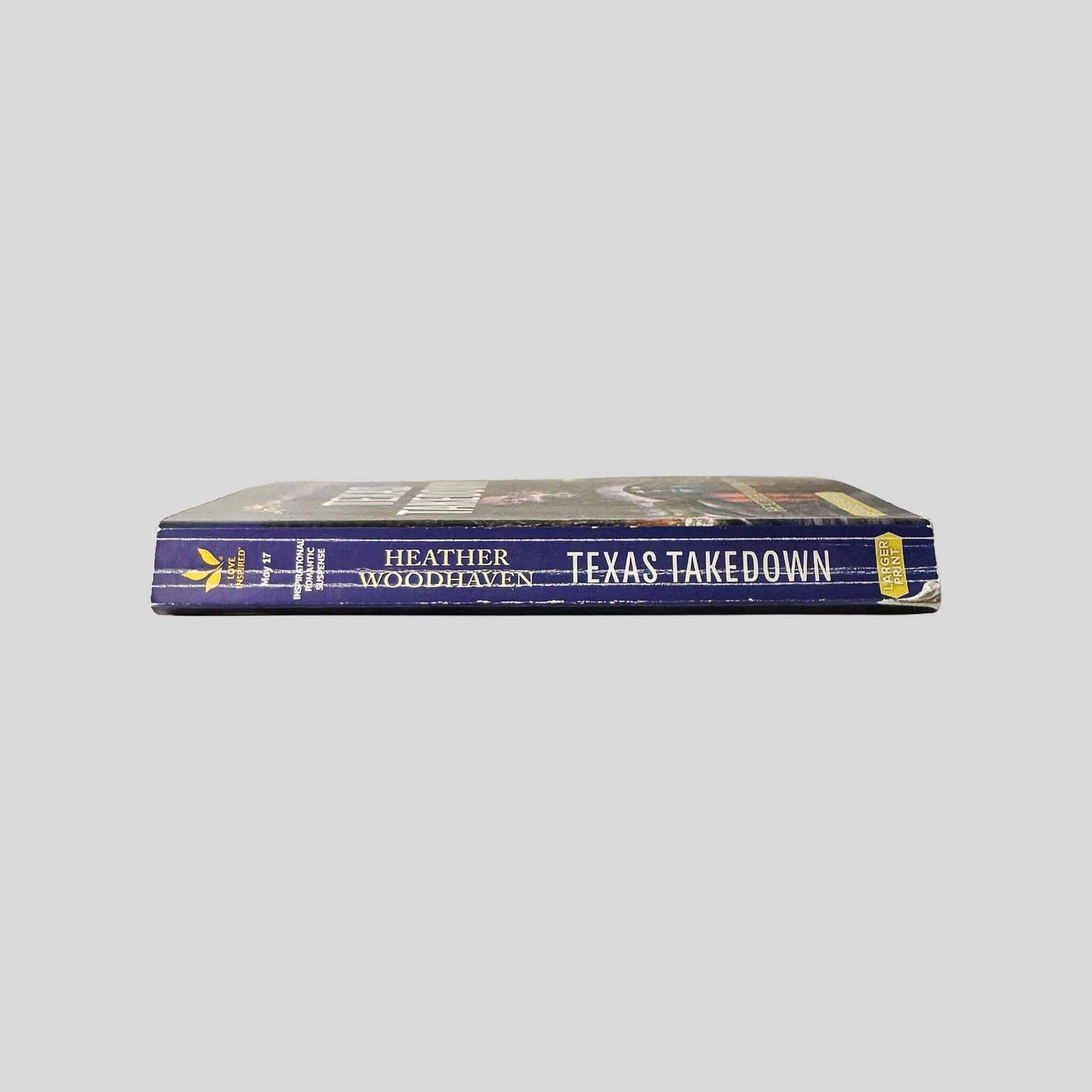 Close-up spine view of Texas Takedown by Heather Woodhaven Large Print centered on a neutral light gray background. Spine shows creasing. Chipping to lower back corner of spine. Pages lay flat.