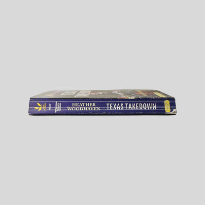 Close-up spine view of Texas Takedown by Heather Woodhaven Large Print centered on a neutral light gray background. Spine shows creasing. Chipping to lower back corner of spine. Pages lay flat.