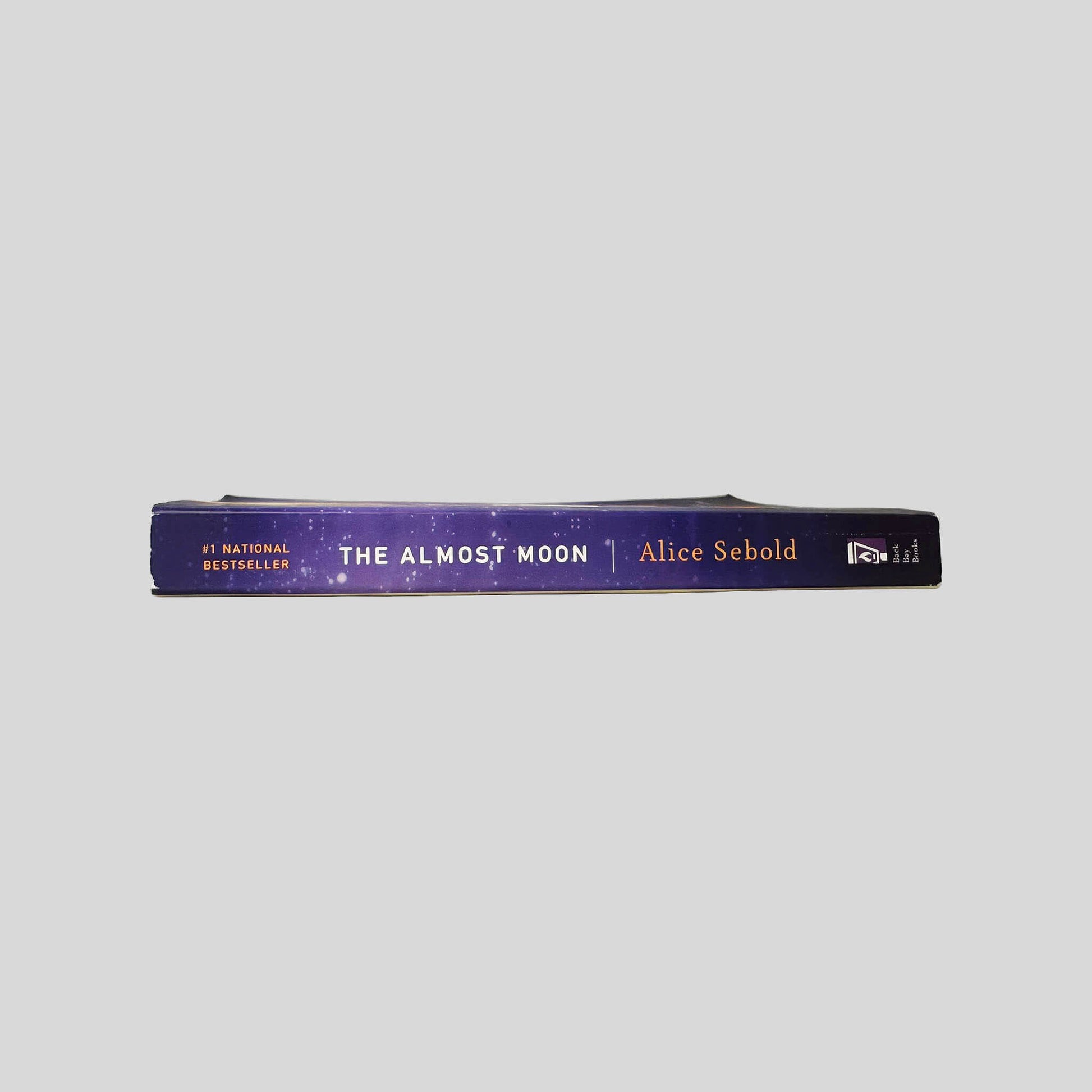 Close-up spine view of The Almost Moon by Alice Sebold centered on a neutral light gray background. Spine is square and shows no creasing. Pages lay flat.