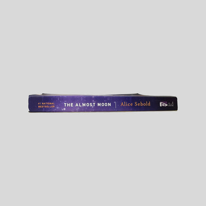 Close-up spine view of The Almost Moon by Alice Sebold centered on a neutral light gray background. Spine is square and shows no creasing. Pages lay flat.