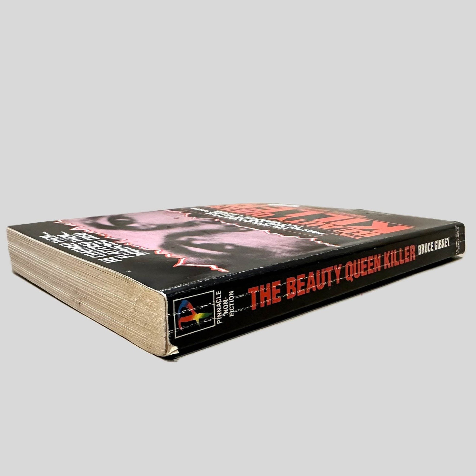 Close-up spine view of The Beauty Queen Killer by Bruce Gibney centered on a neutral light gray background. Spine shows creasing.