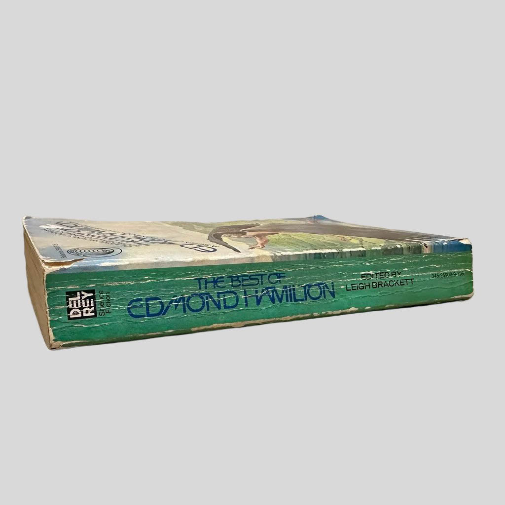 Close-up spine view of The Best of Edmond Hamilton edited by Leigh Brackett centered on a neutral light gray background. Spine is square; shows creasing and chipping to the top and lower end of the spine. Pages lay flat.