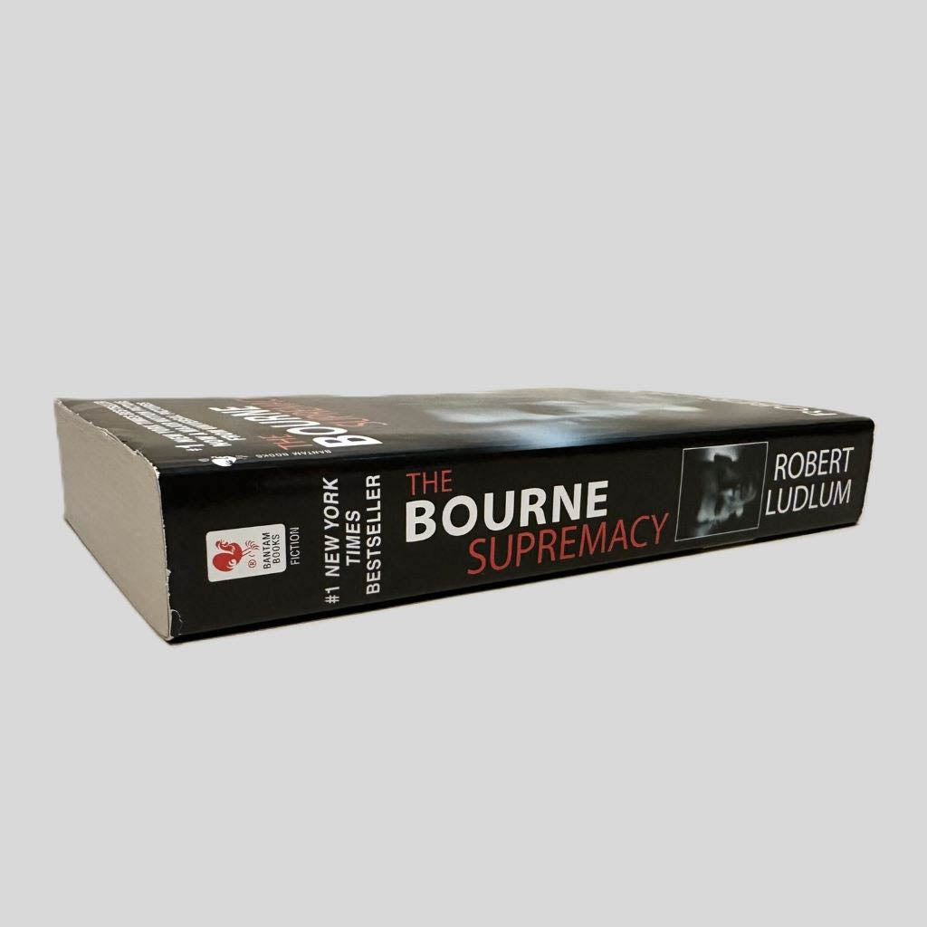 Close-up spine view of The Bourne Supremacy by Robert Ludlum centered on a neutral light gray background. Spine is square and shows minimal creasing. Pages lay flat.