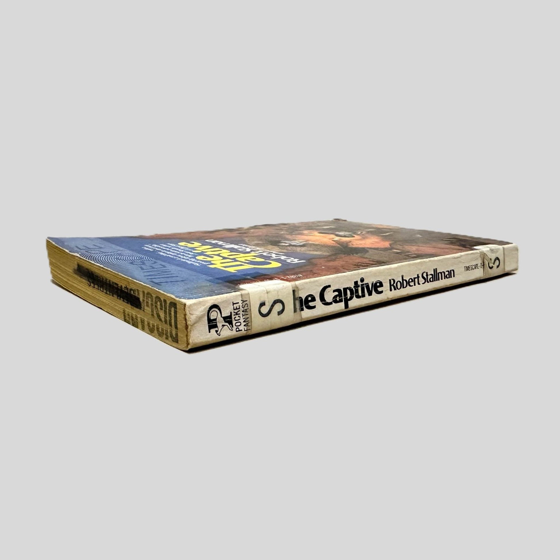 Close-up spine view of The Captive by Robert Stallman centered on a neutral light gray background. Spine shows creasing and bumping. Discard is stamped across the top page edge. The letter "S" is taped once to the top of the spine and again at the bottom for library cataloging. Pages lay flat.