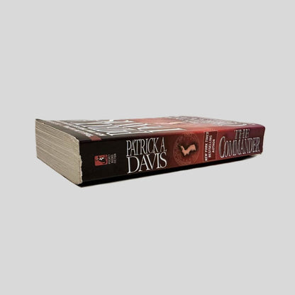 Close-up spine view of The Commander by Patrick A. Davis centered on a neutral light gray background. Spine shows creasing and chipping along the top of the spine. Pages lay flat.