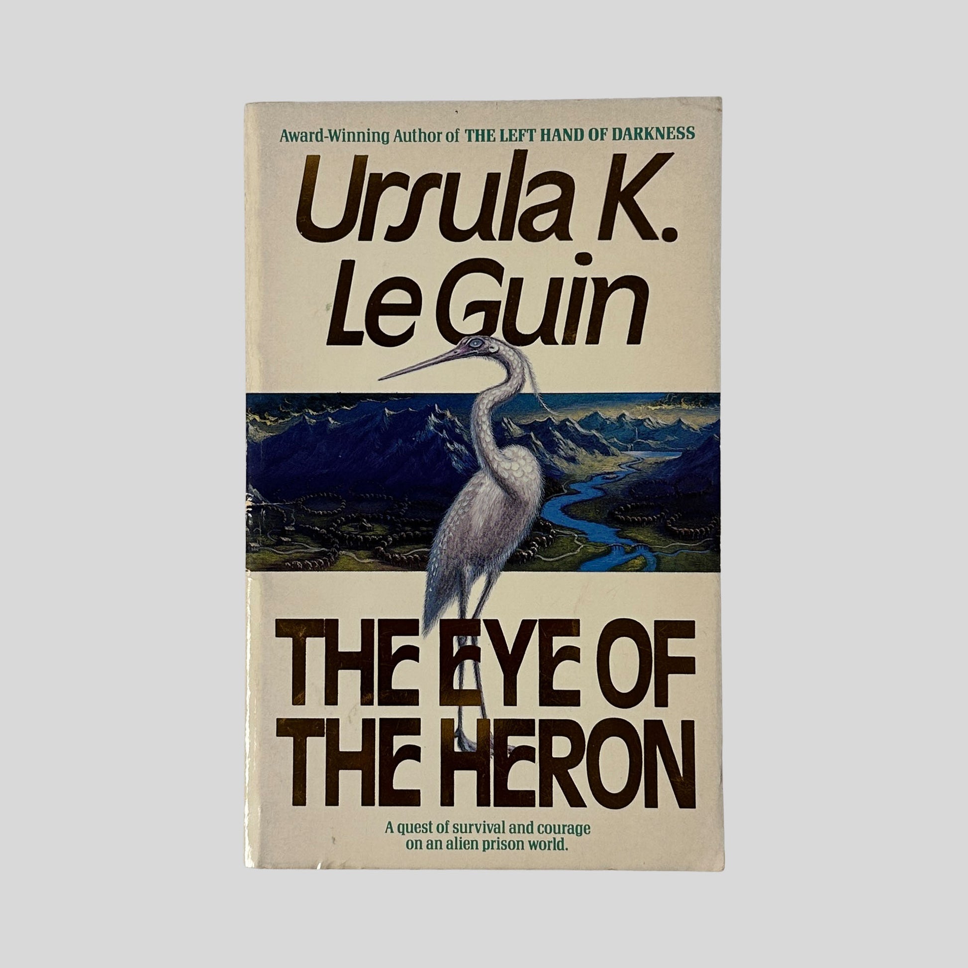 The front cover of the paperback The Eye of the Heron by Ursula K. Le Guin, featuring a large white heron standing before a blue and purple mountainous landscape.