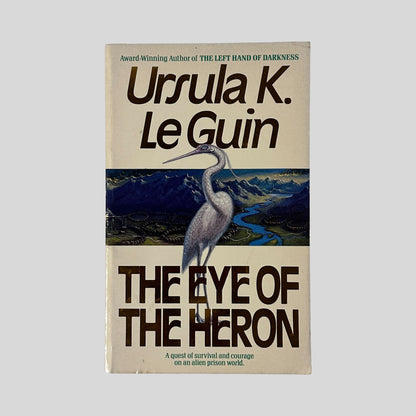 The front cover of the paperback The Eye of the Heron by Ursula K. Le Guin, featuring a large white heron standing before a blue and purple mountainous landscape.
