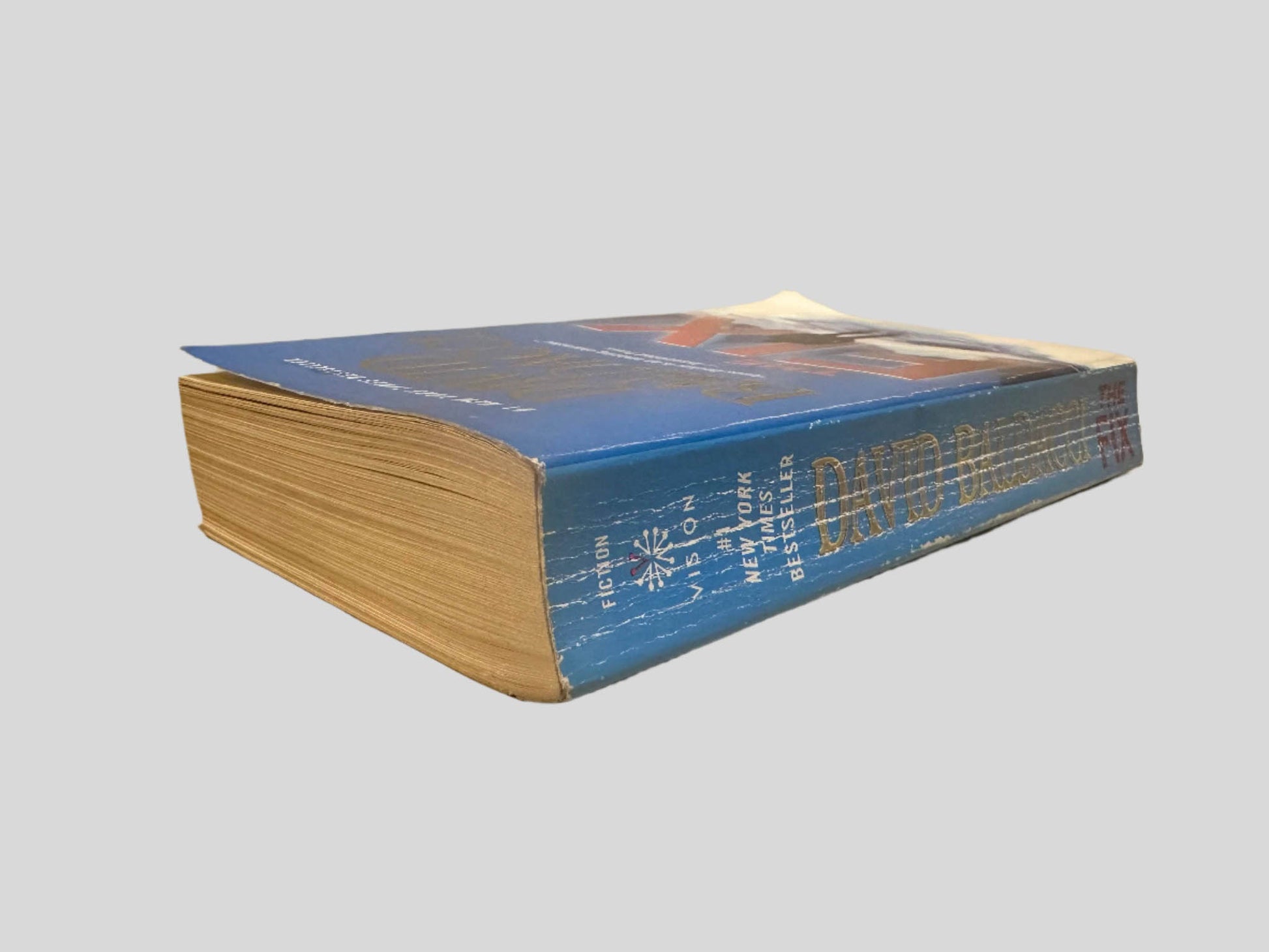 Close-up spine view of The Fix by David Baldacci centered on a neutral light gray background. Spine shows creasing. Pages lay flat.