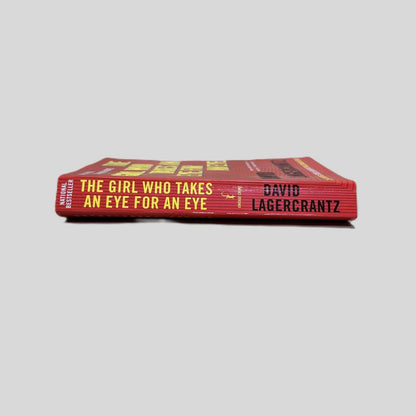 Close-up spine view of The Girl Who Takes an Eye for an Eye by David Lagercrantz centered on a neutral light gray background. Spine shows minimal creasing. Pages lay flat.