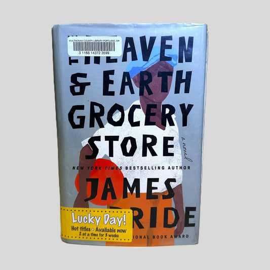 Front cover view of The Heaven and Earth Grocery Store by James McBride centered on a neutral light gray background. Dust jacket is present with mylar cover. Library barcode sticker is present in the top left corner and a "Lucky Day!" library advertisement in the bottom left corner. Cover art is vibrant and intact.