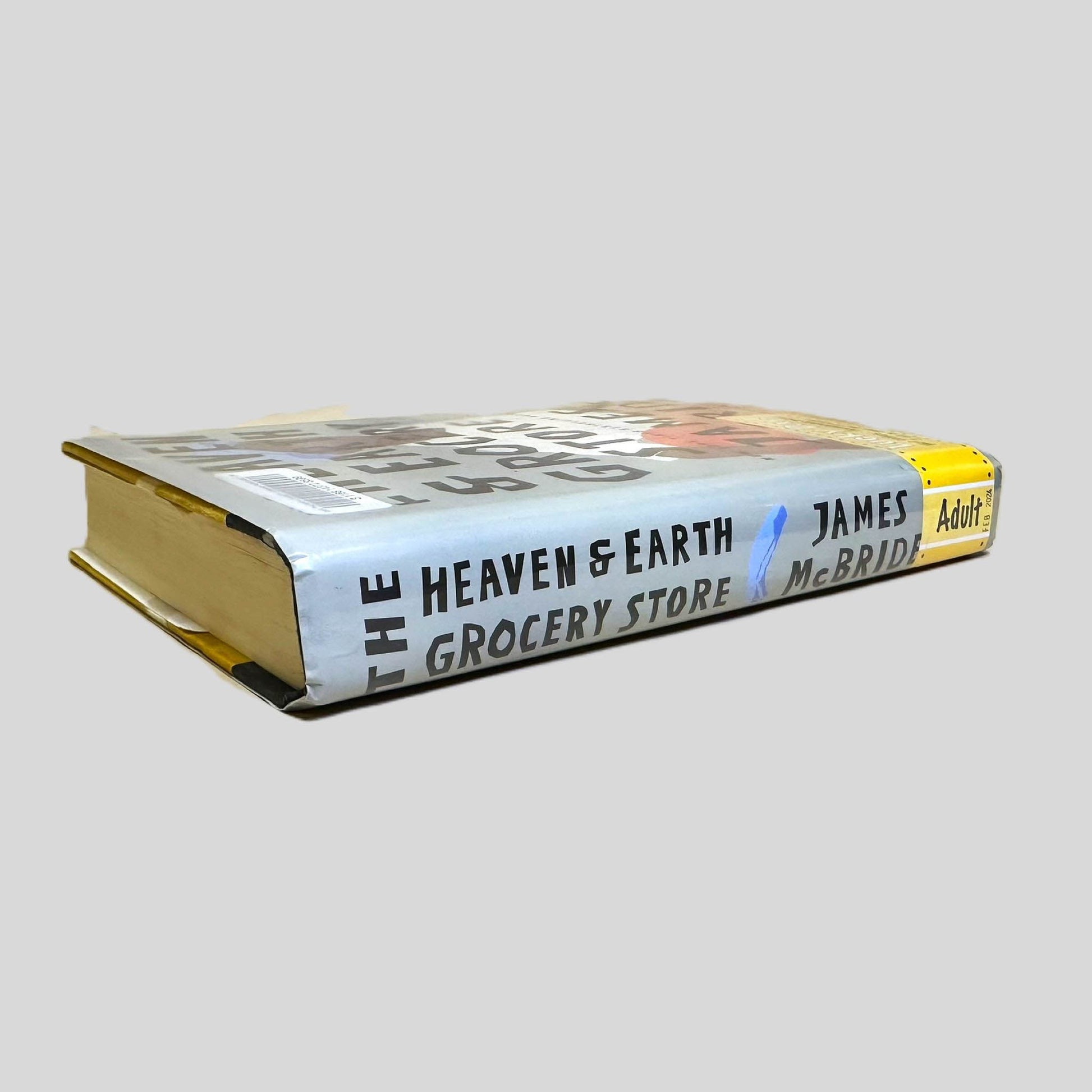 Close-up spine view of The Heaven and Earth Grocery Store by James McBride centered on a neutral light gray background. Spine shows minimal creasing. Pages lay flat.