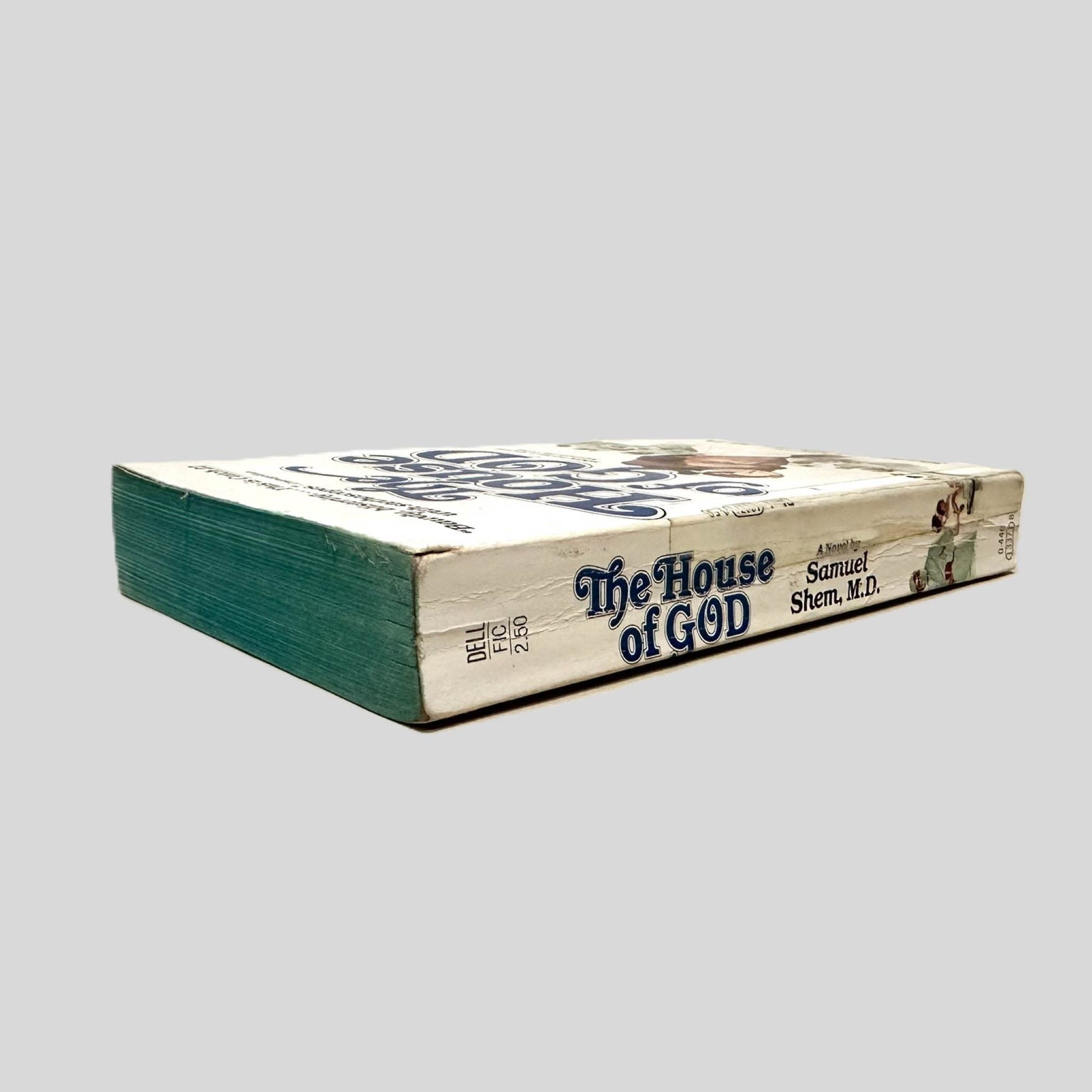 Close-up spine view of The House of God by Samuel Shem, M.D. centered on a neutral light gray background. Spine is square, shows creasing and chipping. Transparent tape is present over the lower part of the spine. Pages lay flat.