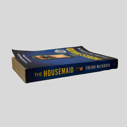 Close-up spine view of The Housemaid by Freida McFadden centered on a neutral light gray background. Spine is square, shows minimal creasing. Pages lay flat.