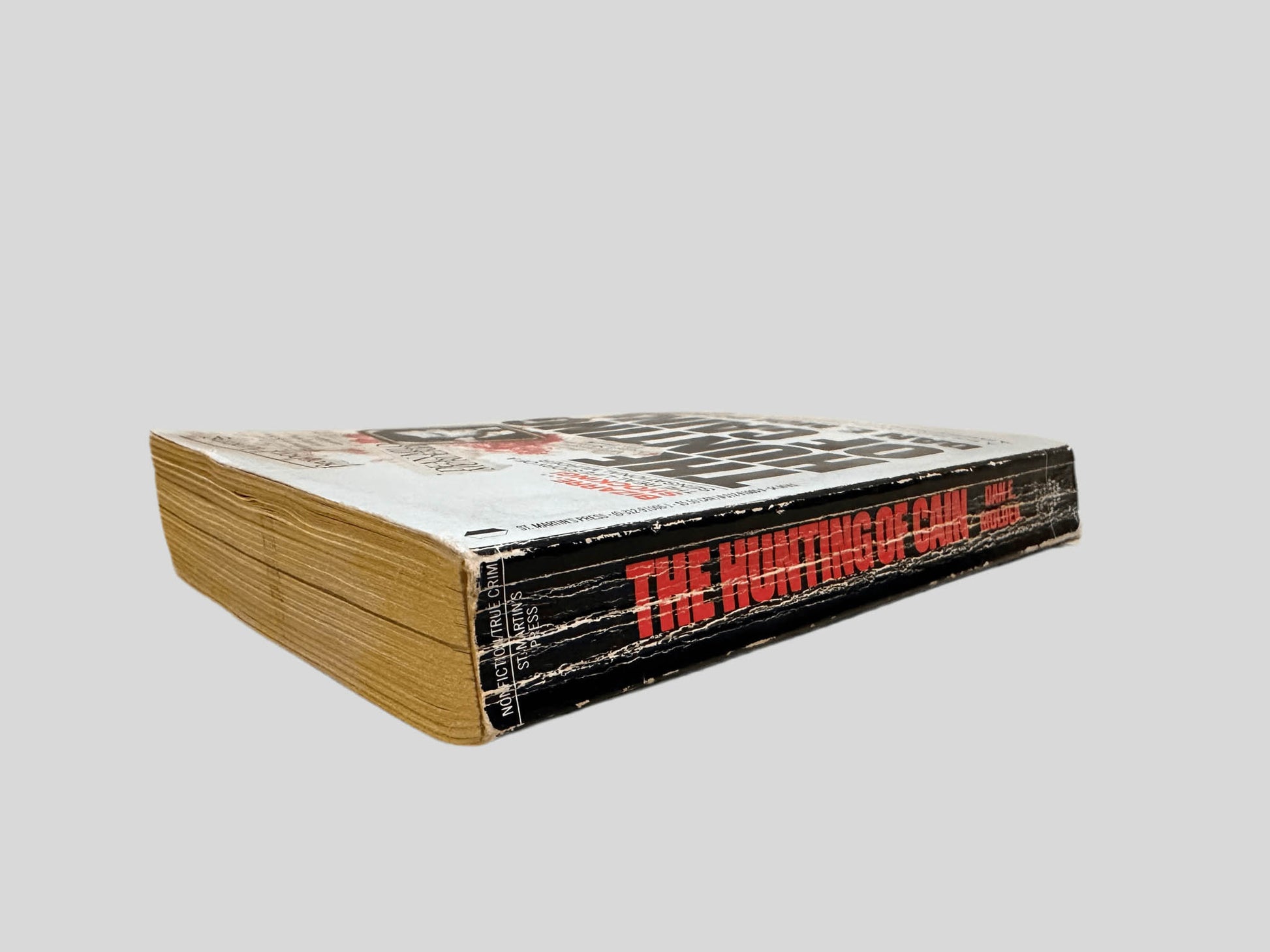 Close-up spine view of The Hunting of Cain by Dan E. Moldea centered on a neutral light gray background. Spine shows creasing and rubbing.