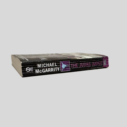 Close-up spine view of The Judas Judge by Michael McGarrity centered on a neutral light gray background. Spine shows creasing.