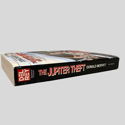 Close-up spine view of The Jupiter Theft by Donald Moffitt centered on a neutral light gray background. Spine is square and shows no creasing. Rubbing marks are present on the edges of spine. Pages lay flat.