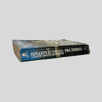 Close-up spine view of The Kingdom by the Sea by Paul Theroux centered on a neutral light gray background. Spine shows creasing and chipping to the top and bottom.  Pages lay flat.