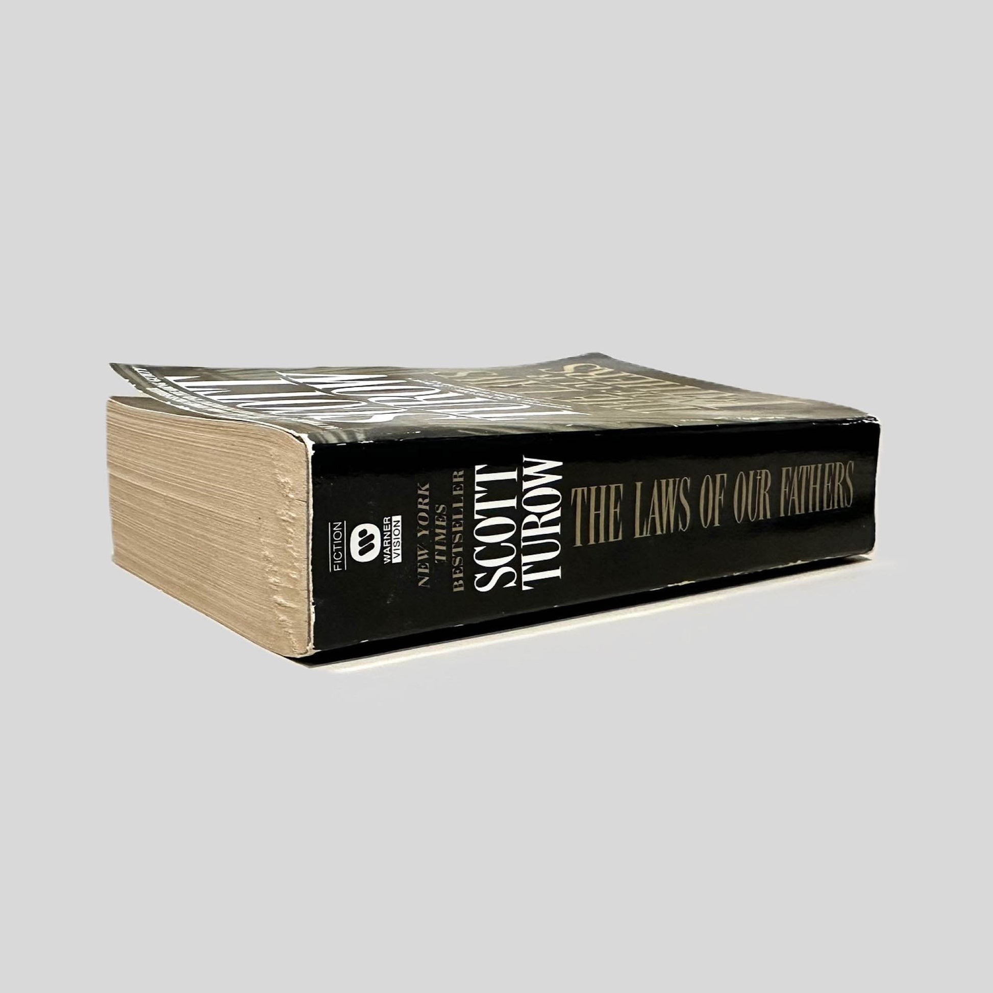 Close-up spine view of The Laws of Our Fathers by Scott Turow centered on a neutral light gray background. Spine is square and shows minor creasing.
