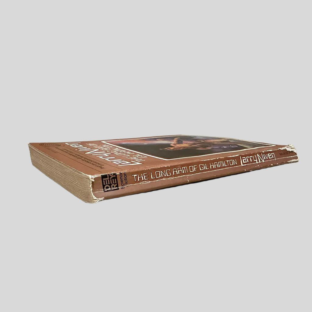Close-up spine view of The Long Arm of Gil Hamilton by Larry Niven centered on a neutral light gray background. Spine shows cocking and creasing. Significant chipping at the bottom edges of the spine. Pages are intact and lay flat.