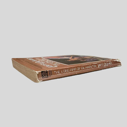 Close-up spine view of The Long Arm of Gil Hamilton by Larry Niven centered on a neutral light gray background. Spine shows cocking and creasing. Significant chipping at the bottom edges of the spine. Pages are intact and lay flat.