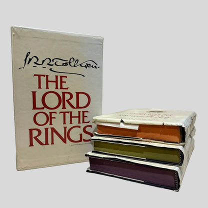 Three-volumes of The Lord of the Rings by J.R.R. Tolkien showing the orange, green, and purple colored page edges, stacked next to the protective outer box. The dust jackets show varying degrees of wear. Pages are tight and lay flat.