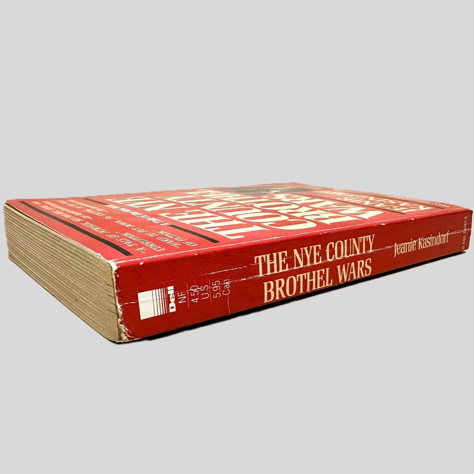 Close-up spine view of The NYE County Brothel Wars by Jeanie Kasindorf centered on a neutral light gray background. Spine shows creasing and chipping.