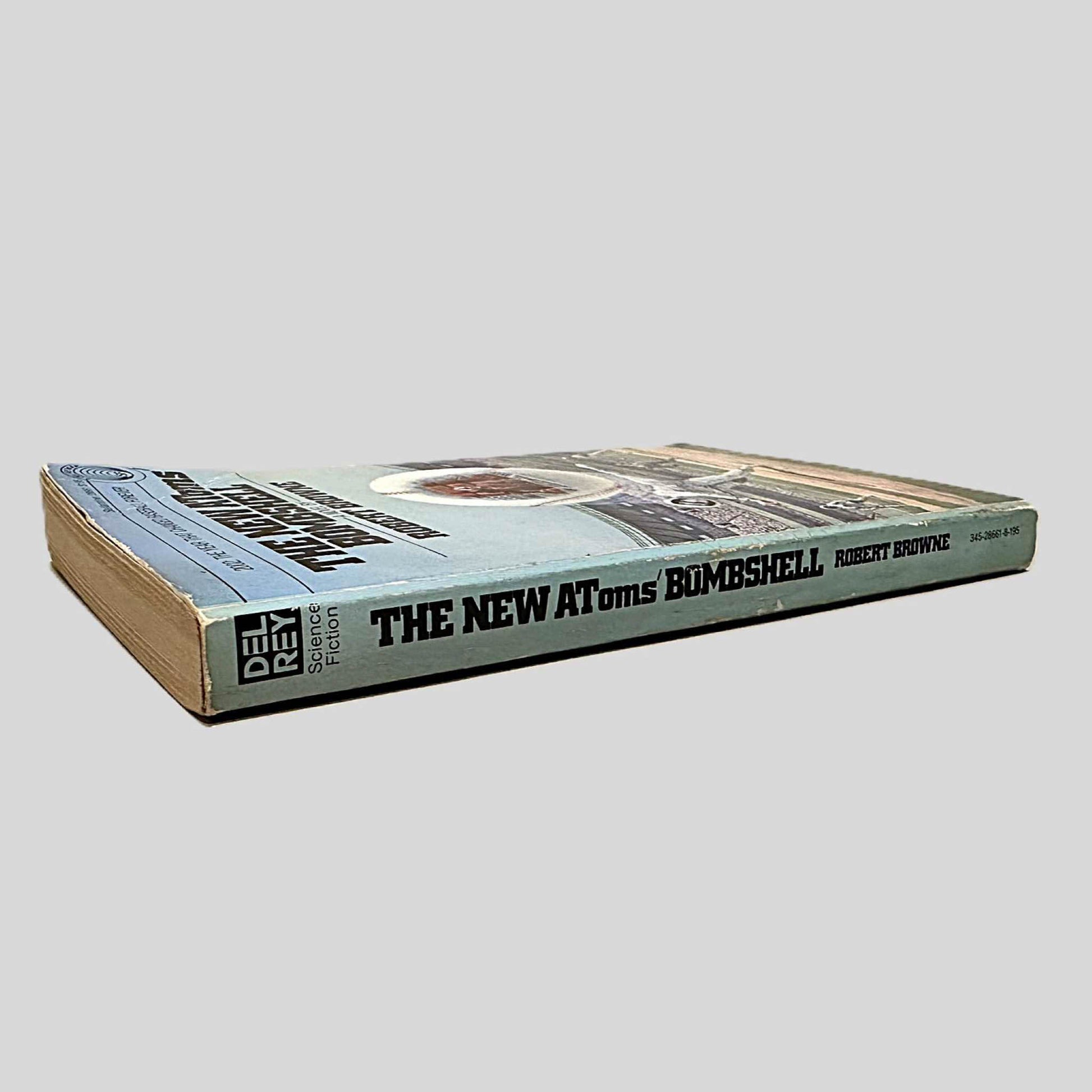 Close-up spine view of The New Atoms' Bombshell by Robert Browne centered on a neutral light gray background. Spine is square with minor chipping and creasing. Pages lay flat.