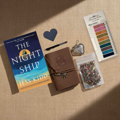 Detailed overhead view of a complete curated collection for 'The Night Ship' novel, laid on a burlap surface. The set features the hardcover book by Jess Kidd, a brown leather-bound journal with compass embossing, an anchor charm cord, black pen, and grey leather heart bookmark. Also included is a clear pouch of loose-leaf herbal tea, a stainless steel tea infuser ball, and a pack of transparent 'Misty Harbor' annotation tabs from Fehmerling Books.