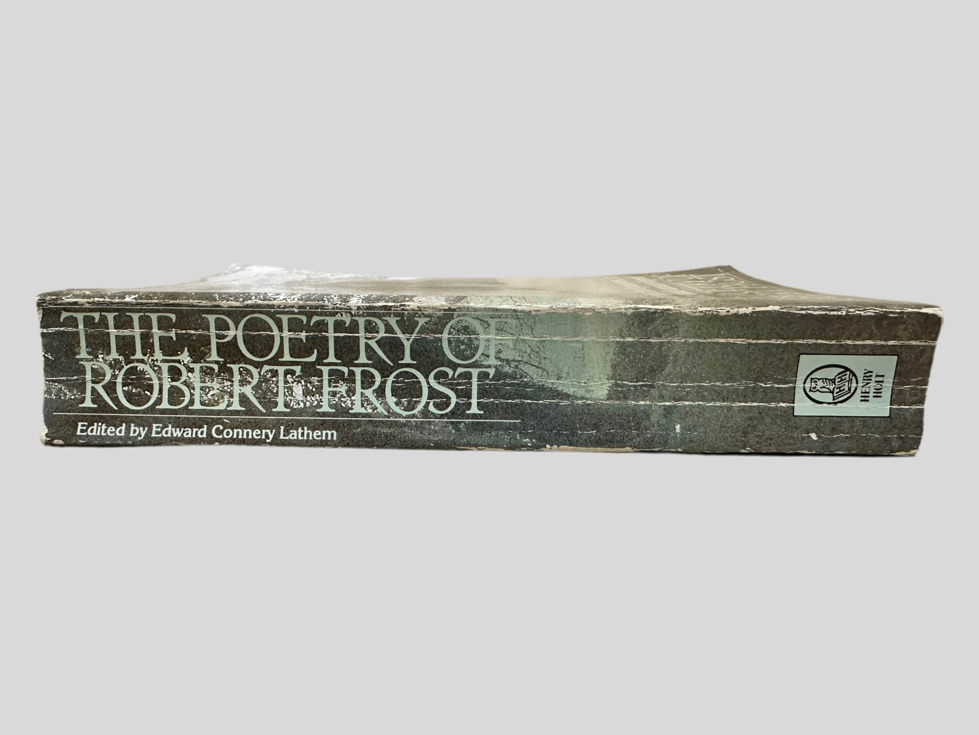 Close-up spine view of The Poetry of Robert Frost: The Collected Poems, Complete and Unabridged centered on a neutral light gray background. Spine shows creasing and chipping. Pages lay flat.