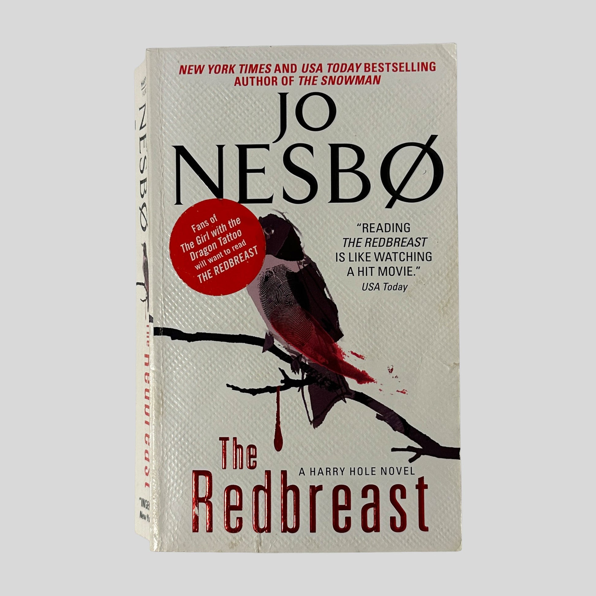 Front cover of the paperback "The Redbreast" by Jo Nesbø. It features a textured white background with a small bird perched on a black branch, with a single red drop of blood dripping from the wood.