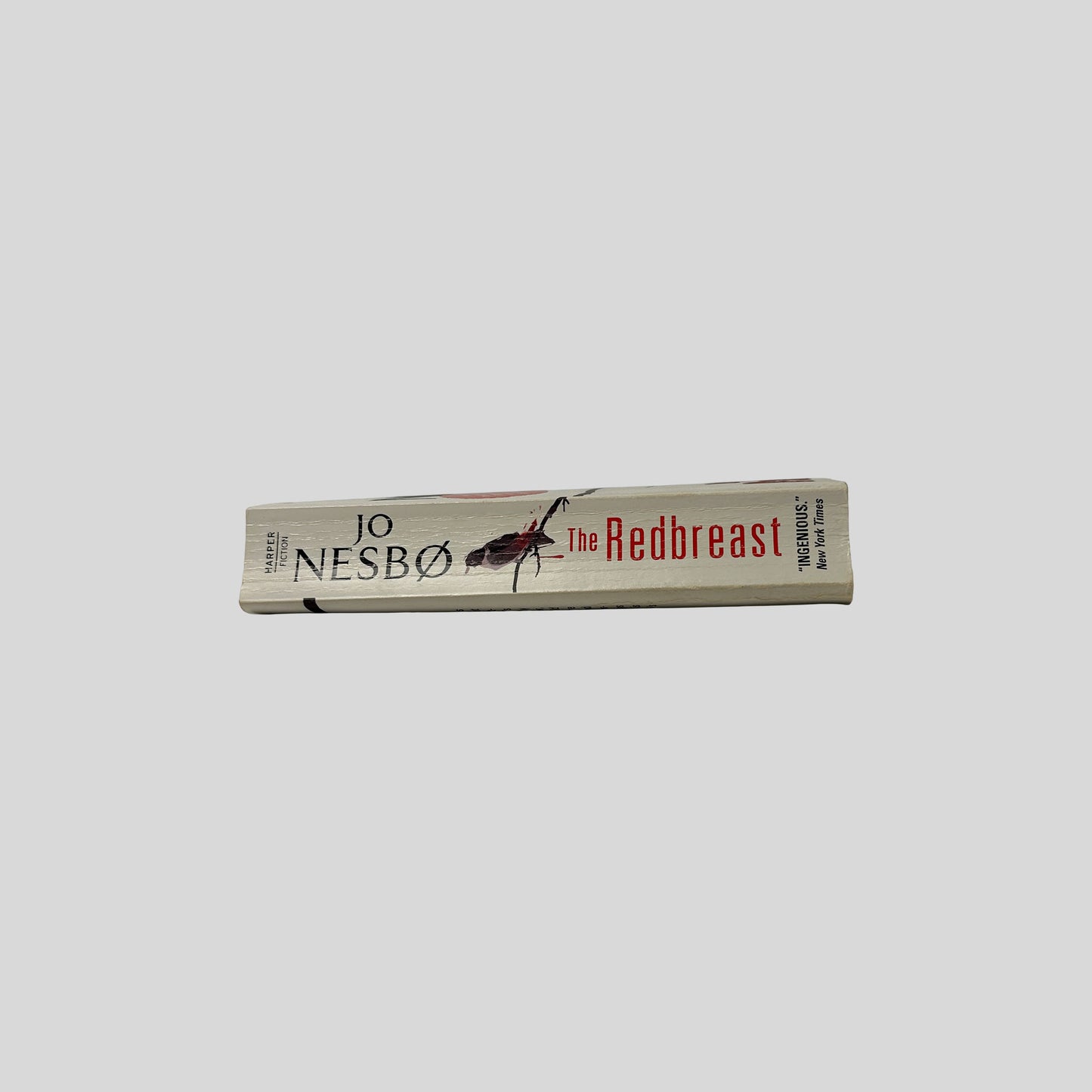 The cream-colored spine of the book "The Redbreast" by Jo Nesbø, featuring the title in red serif font and a small graphic of a small bird perched on a black branch, with a single red drop of blood dripping from the wood.