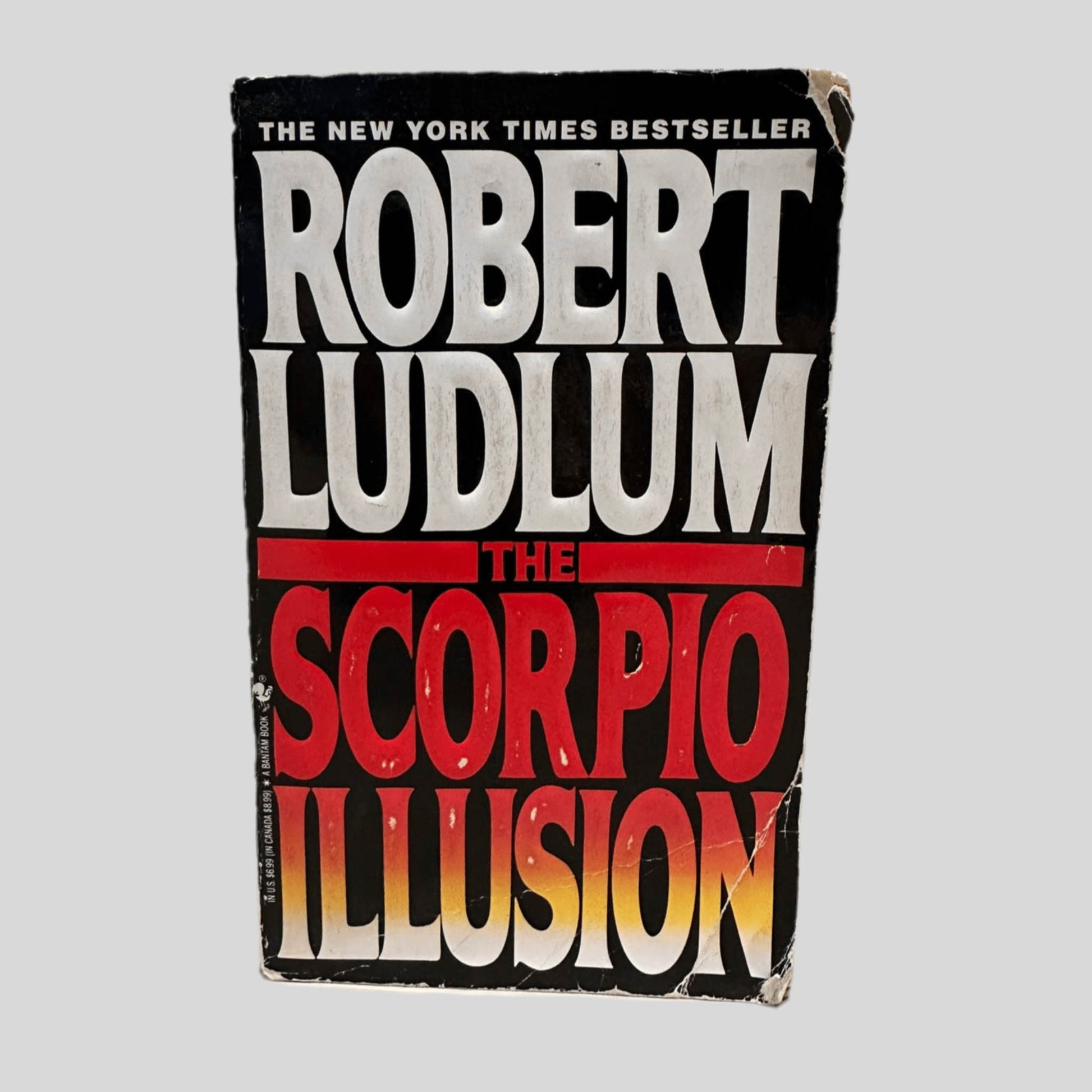 Front cover view of The Scorpio Illusion by Robert Ludlum centered on a neutral light gray background. Chiping present around the edges of the front cover and on the corners. Creasing across the bottom right corner.