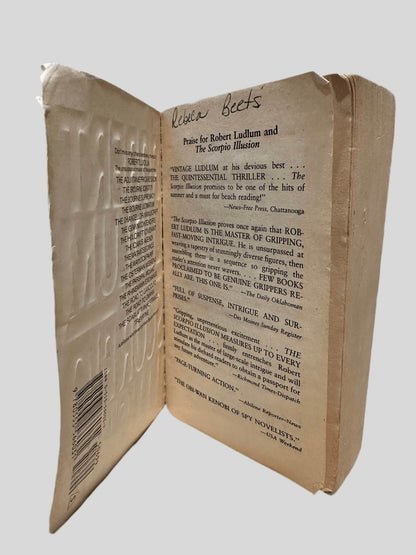 Inside front cover view of The Scorpio Illusion by Robert Ludlum centered on a neutral light gray background. Left hand page has the name "Rebeca Beets" written in black ink at the top.