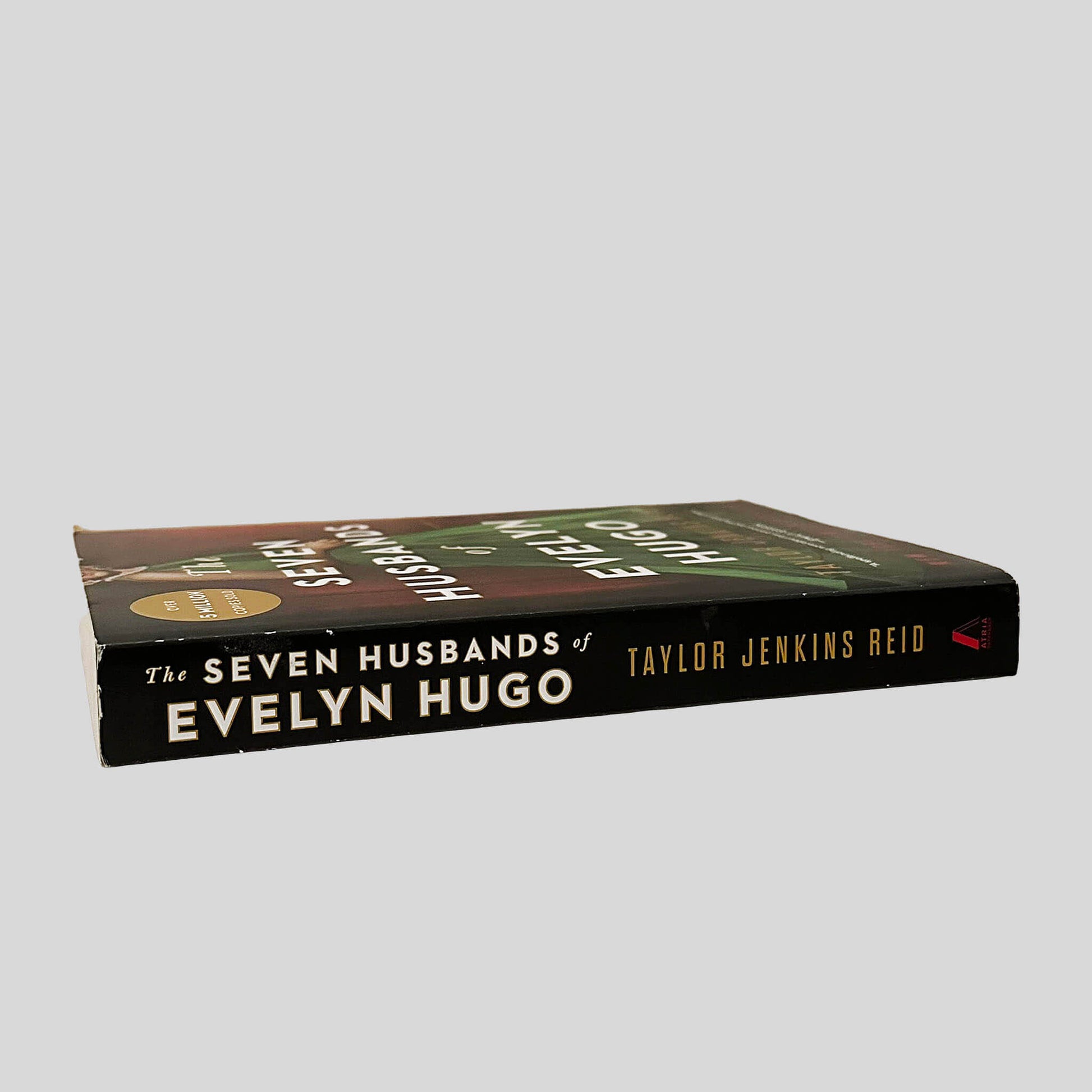Close-up spine view of The Seven Husbands of Evelyn Hugo by Taylor Jenkins Reid centered on a neutral light gray background. Spine is square and shows minimal creasing. Pages lay flat.