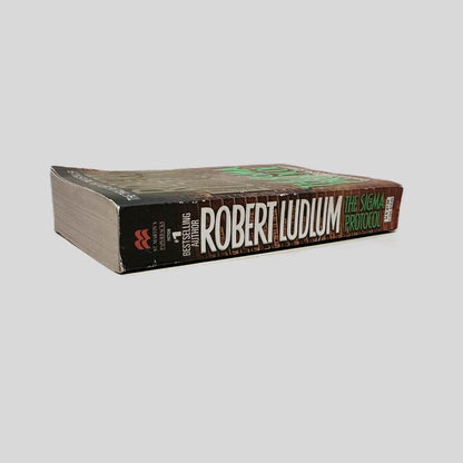 Close-up spine view of The Sigma Protocol by Robert Ludlum centered on a neutral light gray background. Spine is square and shows minimal creasing. Pages lay flat.