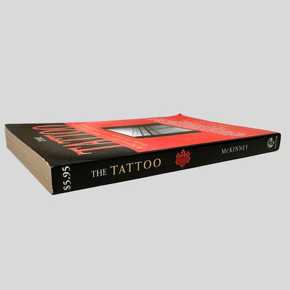 Close-up spine view of The Tattoo by Chris McKinney centered on a neutral light gray background. Spine is square and shows no creasing. Pages lay flat.