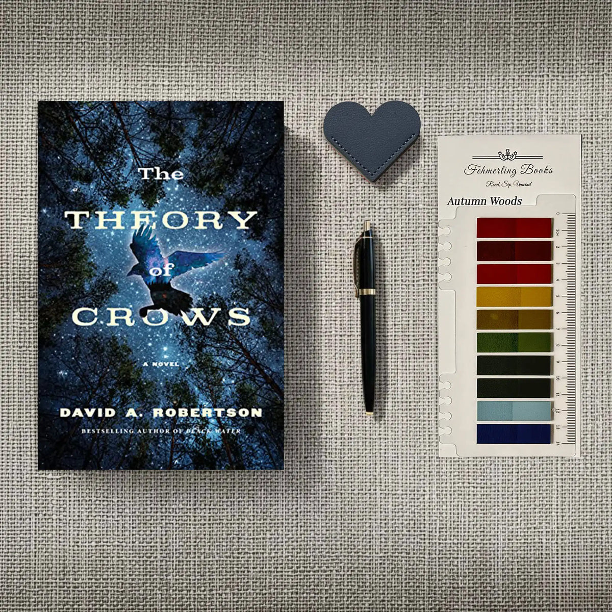 A styled flat-lay featuring the book 'The Theory of Crows' by David A. Robertson, a black pen, a dark blue heart-shaped leather leather bookmark, and a card of 'Autumn Woods' annotation tabs, all arranged on a textured beige fabric background.